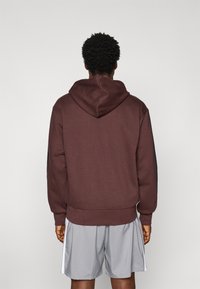 Bordeauxrode hooded sweatshirt met een relaxte pasvorm, ribbelzoom en manchetten, en dubbele zwarte zijpanelen. Draagt met grijze sportshorts.