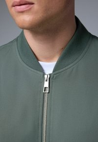 Chaqueta bomber de tela verde apagado, con cuello acanalado y cierre frontal con cremallera y tira de extracción plateada.