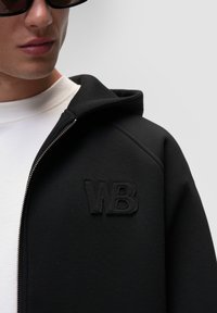 Felpa nera con zip e polsini a coste, caratterizzata da un logo ricamato "WB" sul petto, indossata sopra una t-shirt bianca. Texture liscia.