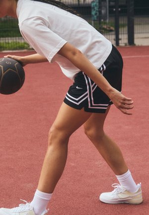 Personne dribble un ballon de basket noir sur un terrain extérieur rouge, portant un short noir, un t-shirt blanc, des chaussettes blanches et des baskets blanches.
