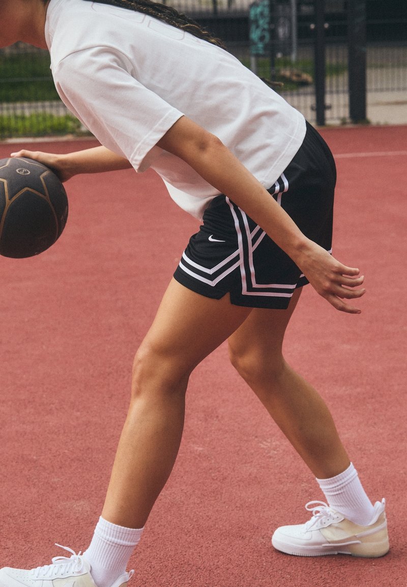 Personne dribble un ballon de basket noir sur un terrain extérieur rouge, portant un short noir, un t-shirt blanc, des chaussettes blanches et des baskets blanches.