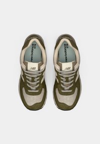 Maslinasto zelena i bež New Balance 574 tenisice s teksturiranim gornjim dijelom, okruglim vrhom i sivim vezicama. Sadrži logo na jeziku i sa strane.