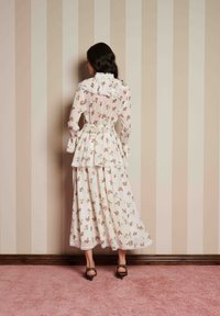 Μακρυμάνικο φόρεμα με λουλουδάτο σχέδιο σε απαλό ροζ, με ριχτό γιακά και φούστα σε στρώσεις. Φορεμένο με περίτεχνα μαύρα τακούνια. Ο τοίχος έχει κατακόρυφες ρίγες.