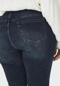 Jeans de mezclilla azul oscuro con un diseño ajustado, que cuentan con bolsillos traseros y un desgastado sutil. La textura es suave con un ligero brillo.