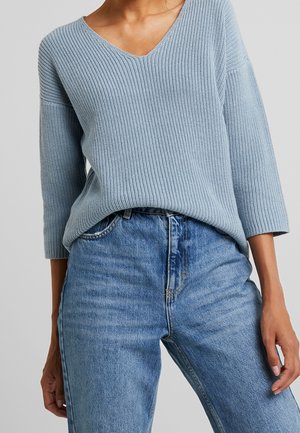 Femme portant un pull en tricot côtelé bleu clair à manches trois-quarts, rentré dans un jean bleu taille haute.