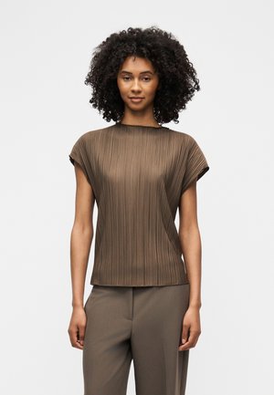 Nainen, jolla on kiharat hiukset, pukeutuneena ruskeaan laskostettuun lyhythihaiseen puseroon ja vastaaviin ruskeisiin housuihin, seisoo yksinkertaista valkoista taustaa vasten.