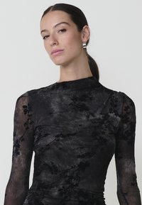 Bluză neagră semi-transparentă cu textură florală, mâneci lungi și decolteu înalt. Prezintă detalii plisate și o suprafață netedă, ușor lucioasă.