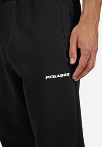 Nærbillede af en hånd, der hviler ved sorte joggingbukser med frontlommer og "PEGADOR" trykt i hvidt på låret.