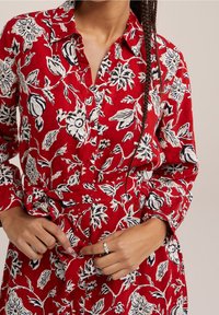 Röd blommig skjortklänning med vita och svarta mönster. Har krage, knappstängning framtill och ett midjeband.