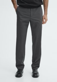 Pantalon gris à jambes droites en tissu tissé lisse. Dispose de poches latérales et d'une taille traditionnelle. Porté avec des chaussures noires.
