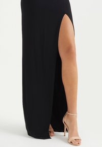 En kvinnas ben synligt genom en hög skena i en lång svart klänning, bärande nakna högklackade sandaler med ankelrem på ljus bakgrund.