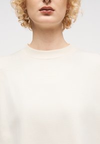 Ξανθό άτομο με σγουρά μαλλιά, φοράει μικρούς κρίκους σκουλαρίκια και ένα εκρού πουλόβερ με στρογγυλή λαιμόκοψη, με διακριτικό ανάγλυφο κείμενο.