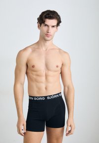 Schwarze Boxer-Briefs aus weichem Stoff mit engem Sitz. Mit einem weißen Logo-Bund und einem klassischen Design. Glatte Textur, Oberschenkel-lange.