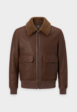 Homme portant une veste en cuir marron avec col en fourrure, polo bleu marine, jean gris et chaussures marron, debout sur fond blanc.