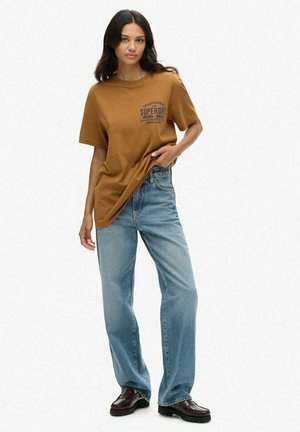 Brun bomulds t-shirt med korte ærmer og grafisk tekst, parret med lyseblå posede denim jeans og mørke polerede sko.