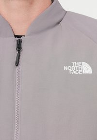 Ανοιχτό γκρι μπουφάν με όρθιο γιακά, διαθέτει μαύρο φερμουάρ και κεντημένο λευκό λογότυπο. Το υλικό φαίνεται λείο με διακριτική υφή.