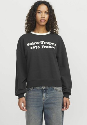 Felpa nera con testo bianco "Saint-Tropez 1970 Francia", collo rotondo, polsini a costine e vestibilità comoda. Indossata con jeans in denim azzurro.