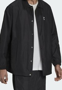 Svart lättviktsjacka med tryckknappar, elastiska ärmslut och sidofickor. Har en liten vit logotyp på vänster bröst.