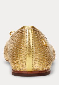 Vävd gyllen platt sko med metallisk rem vid hälen och liten gyllen spänne detaljer på sidan, visad från baksidan.