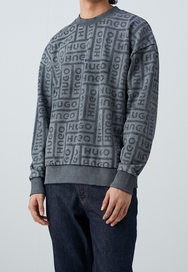 Sudadera gris con un patrón repetido de logotipos, cuello redondo, mangas largas, puños y dobladillo acanalados, y un corte holgado. Combinada con vaqueros oscuros.