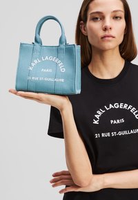 Borsa a tracolla in tela blu chiaro con manici corti, caratterizzata da un testo stampato bianco: "KARL LAGERFELD PARIS 21 RUE ST-GUILLAUME." Forma rettangolare compatta.