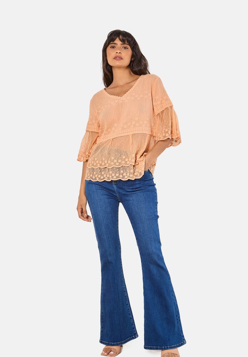 Pfirsichfarbene durchsichtige Bluse mit gewelltem Saum und rüschierten kurzen Ärmeln, kombiniert mit dunkelblauen Schlagjeans. Weiche Stofftextur, lässige Passform.