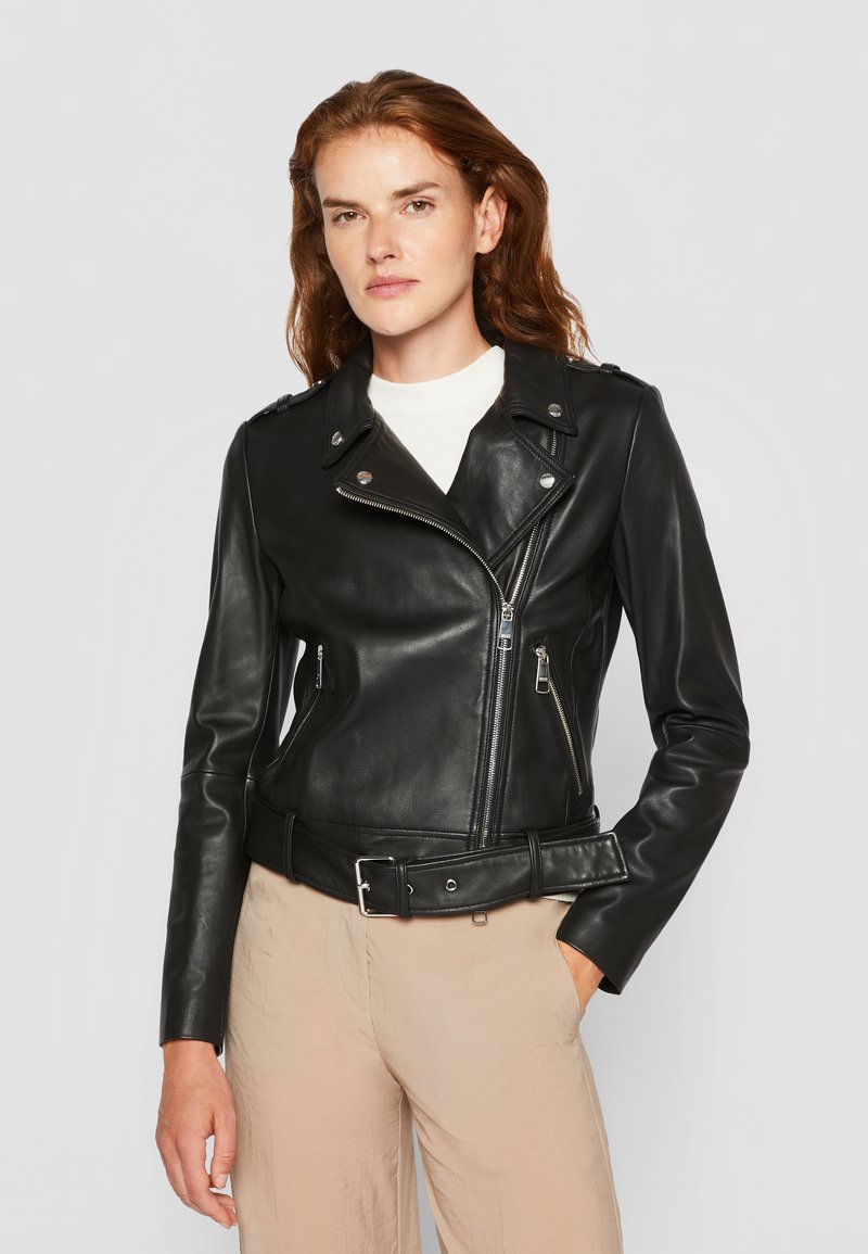 Giacca da motociclista in pelle nera con zip asimmetrica, accessori in argento e colletto a scatto; presenta una vita con cinturino e un design aderente.