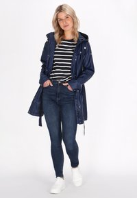 Modra vodoodporna jakna s kapuco in gumbastimi zapirali, nosena nad črno-belimi črtastimi majicami, v paru z temnimi kavbojkami in belimi supergami.