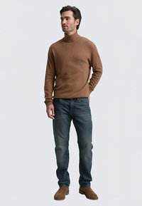 Jersey de cuello vuelto marrón de punto, vaqueros azules y botas de ante color tostado, usados por un modelo masculino de pie frente a un fondo neutro.