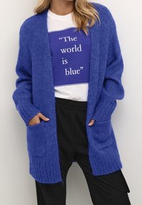 Cardigan azul de punto con textura de canalé, de frente abierto y con bolsillos. Llevado sobre una camiseta gráfica azul y blanca a juego y pantalones negros.