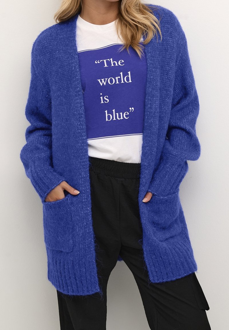 Cardigan azul de punto con textura de canalé, de frente abierto y con bolsillos. Llevado sobre una camiseta gráfica azul y blanca a juego y pantalones negros.