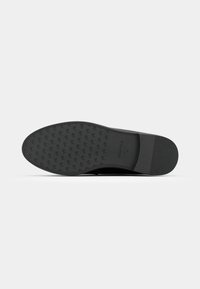 Semelle d'une chaussure habillée noire avec un petit motif de logo texturé, une zone de talon lisse et "ANNA FIELD 37" embossé près de l'arche.