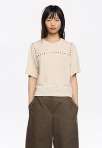 Camisa corta beige con cuello redondo y detalles de tachuelas en el pecho. Combinada con pantalones sueltos verde oliva. Textura suave.