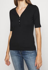 Svart ribbad kortärmad topp med V-ringning och knäppning med tre knappar, tillverkad av ett elastiskt material för en figurnära silhuett.