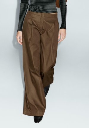 Person wearing high-waisted, wide-leg brown pants with front pleats, paired with a black and gray striped long-sleeve top and black pointed shoes.