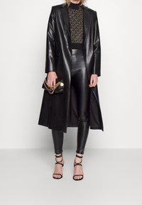 Cappotto lungo in pelle nera su un top nero aderente e leggings. Gli accessori includono una piccola borsa nera con una tracolla in catena dorata e sandali con cinturini.