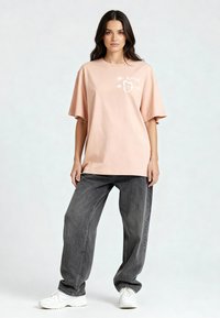 T-shirt oversize rose clair avec le texte et le graphique "Mi Amor" en blanc. Porté avec un jean baggy gris foncé et des baskets blanches. Texture en coton lisse.