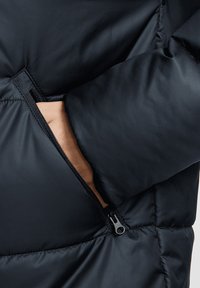 Schwarze Pufferjacke mit kontrastierender brauner Fütterung. Verfügt über eine mit Reißverschluss verschließbare Tasche auf der linken Seite und eine gesteppten Textur.