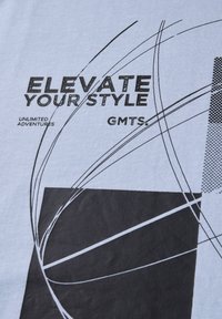 T-shirt graphique bleu clair avec un texte noir et des formes abstraites, y compris des lignes et des blocs. Le texte indique "ÉLEVEZ VOTRE STYLE" et "AVENTURES ILLIMITÉES."