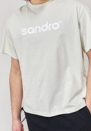 Man draagt een lichtbeige T-shirt met het woord "sandro" in grote witte letters op de borst en een donkere broek.
