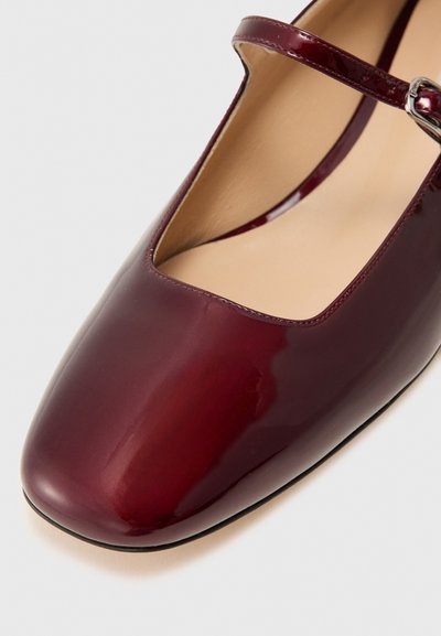 Sapatilha Mary Jane de couro vernice burgundy, com bico quadrado e uma tira fina ajustável com fivela prateada. Superfície interior lisa.