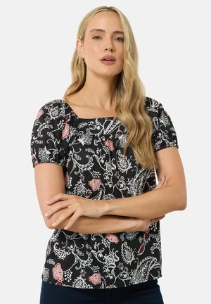 Lyshåret kvinde med langt hår iført sort bluse med korte ærmer og hvide og røde blomsterprint, armene krydset over brystet.