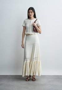 Kobieta stoi, ubrana w kremową, zapinaną na guziki bluzkę z krótkim rękawem oraz długą plisowaną spódnicę, trzyma małą brązową torebkę na ramię i nosi czarne sandały na obcasie.