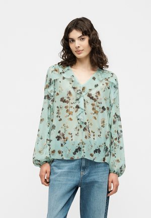Kvinde med krøllet hår iført en lyseblå blomsterprintet gennemsigtig bluse med flæser og lange ærmer kombineret med blå jeans.