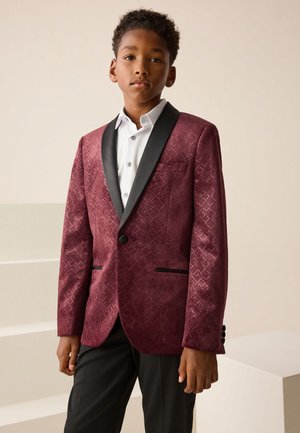Junge trägt einen gemusterten burgunderfarbenen Blazer mit schwarzen Revers, ein weißes Hemd und schwarze Hosen und steht vor einem einfachen Hintergrund.