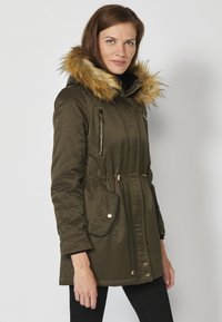 Olivový parka s kapucňou z umelého kožúšku, so zipsom na predných vreckách, sťahovacím pásom a čiernymi akcentmi. Hladká, odolná tkanina s lesklými detalami.