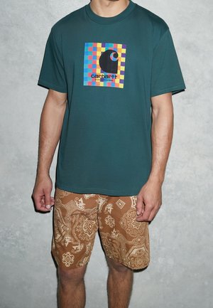 Homme portant un t-shirt Carhartt bleu sarcelle avec un logo à carreaux colorés et un short marron à motifs, debout devant un mur gris texturé.
