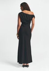 Juoda suknelė be petnešų, pagaminta iš lygios medžiagos, su drapiruotėmis ir ilga, prigludusia siluetu. Derinama su juodomis aukštakulnėmis avalynėmis.