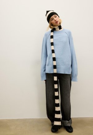 Pull oversize bleu clair, écharpe rayée noir et blanc, jean gris foncé et chaussures noires. Textures simples et couleurs contrastées.