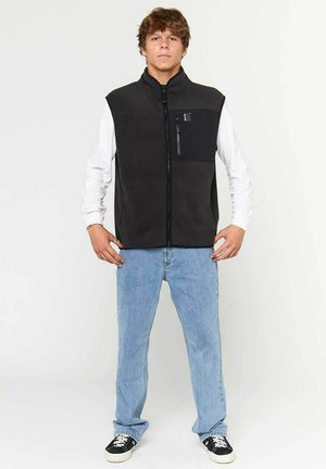 Black fleece vest with a high collar, front zip closure, and a left chest zip pocket. Worn over a white long-sleeve shirt and blue jeans.